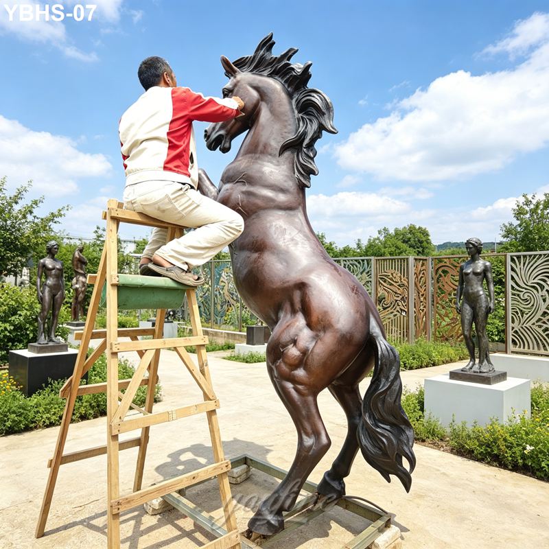 An artist standing on a wooden ladder, working on the details of a large bronze rearing horse sculpture in an outdoor art setting.