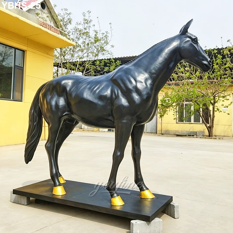 A life-size black bronze horse sculpture with gold accents on hooves, placed outdoors on a sturdy pedestal. Yellow building in the background.