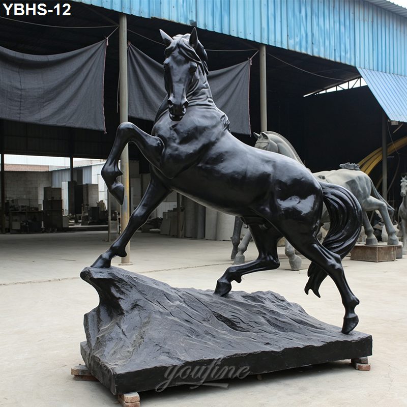 Majestic black bronze horse sculpture rearing on a textured base, displayed in an industrial outdoor setting.