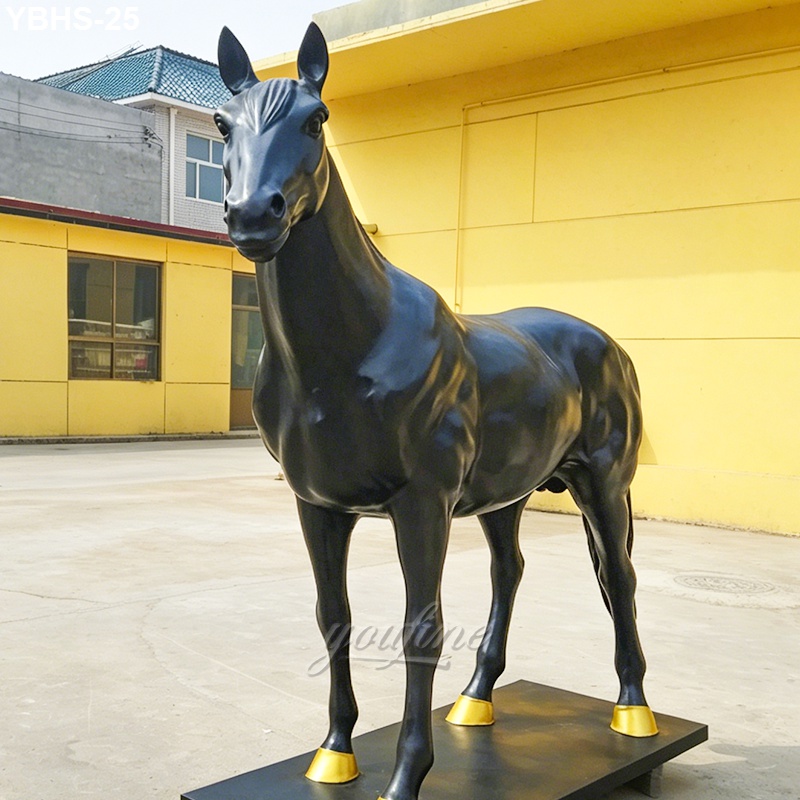 A detailed black bronze horse statue with golden hooves standing on a base, placed outdoors near a yellow building.