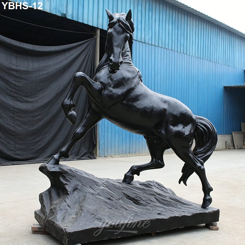 A black bronze horse sculpture rearing on a rugged base, displayed outdoors near a blue structure, symbolizing strength and elegance.