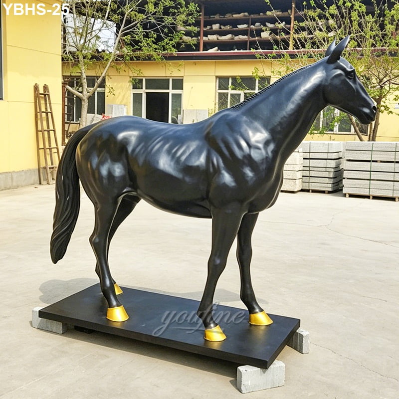 Glossy black bronze horse statue with gold hooves standing on a platform in an outdoor setting with trees and industrial background.
