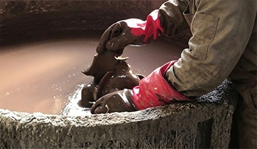 Worker wearing red gloves operates the bronze casting process, handling a mold immersed in liquid bronze within a large vat.