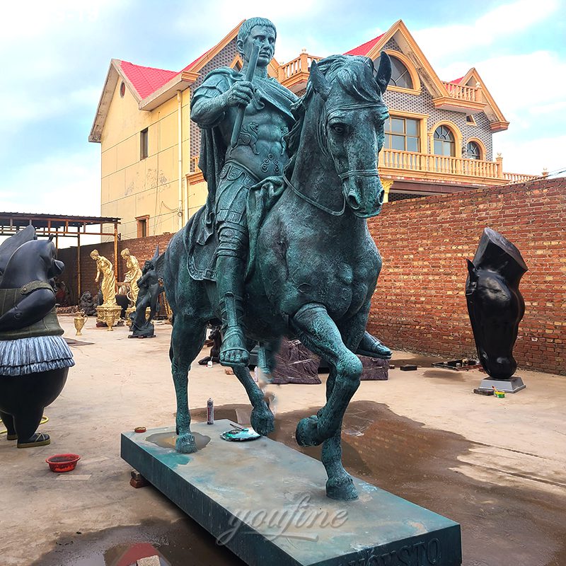 A detailed bronze equestrian statue featuring a warrior on horseback, set outdoors in a workshop surrounded by sculptures and building structures.