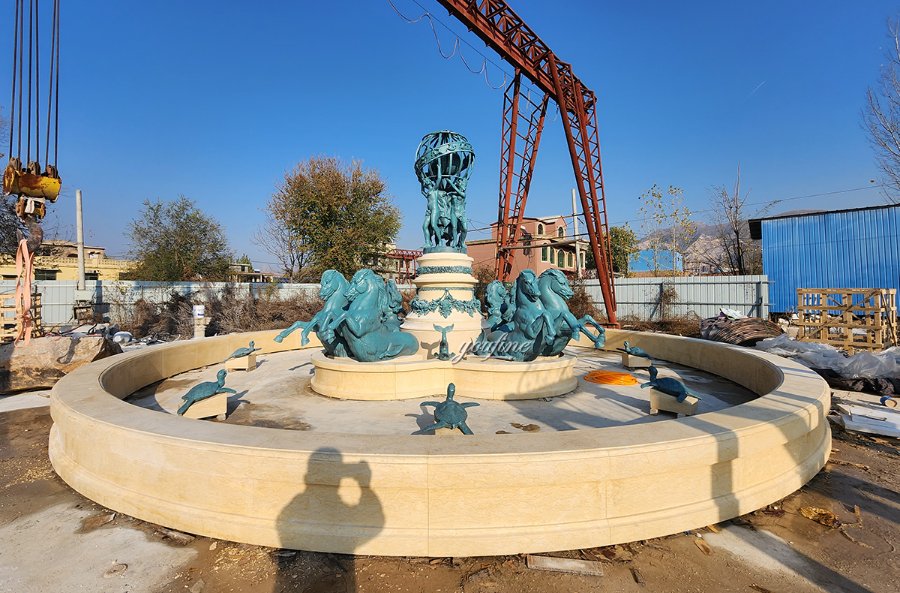 Elaborate bronze fountain sculpture featuring turtles and human figures in a circular basin during installation at an outdoor site.