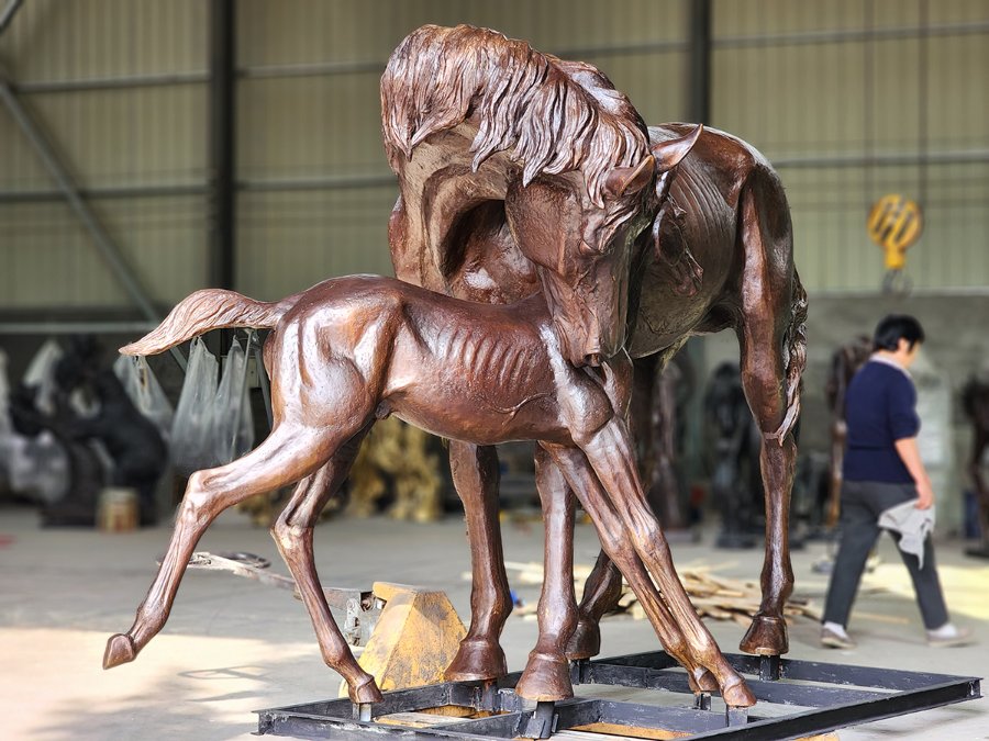 A life-sized bronze sculpture of a horse and its foal in a tender pose, displayed in a workshop setting.