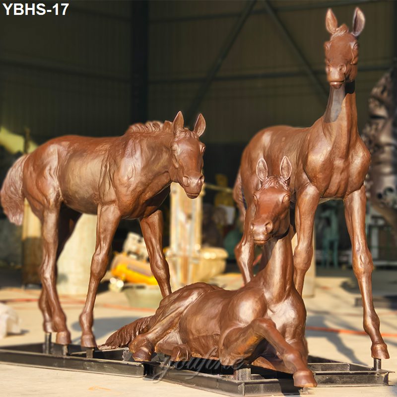 Bronze sculpture of a horse family featuring a standing horse, a foal, and a reclining horse in an indoor workshop setting.