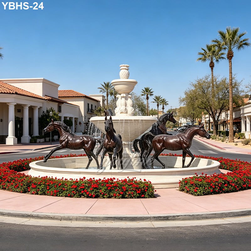 Bronze horse statues surrounding a large fountain in a circular landscaped feature with red flowers and palm trees in a sunny commercial area.