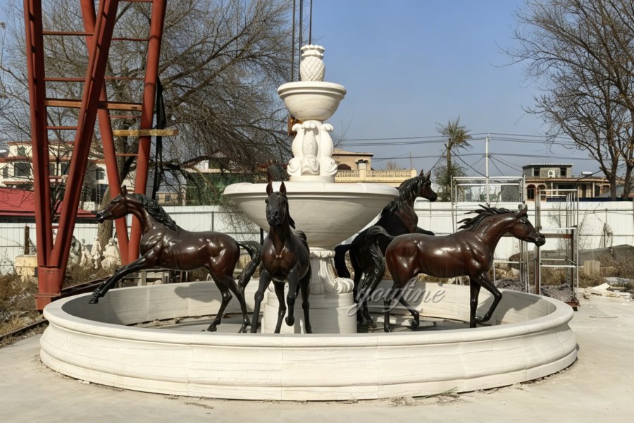 A decorative fountain featuring dynamic bronze horse sculptures encircling a white stone basin with an ornate centerpiece outdoors.