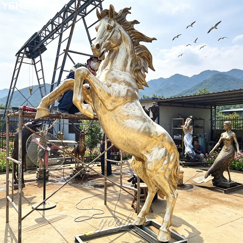 An artist polishes a golden-colored rearing bronze horse sculpture in an outdoor workshop with surrounding statues and mountains in the background.