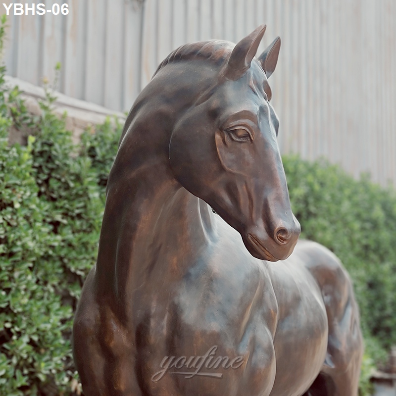 Close-up of an elegant bronze horse sculpture featuring lifelike anatomical details with a natural backdrop of greenery and wooden fencing.