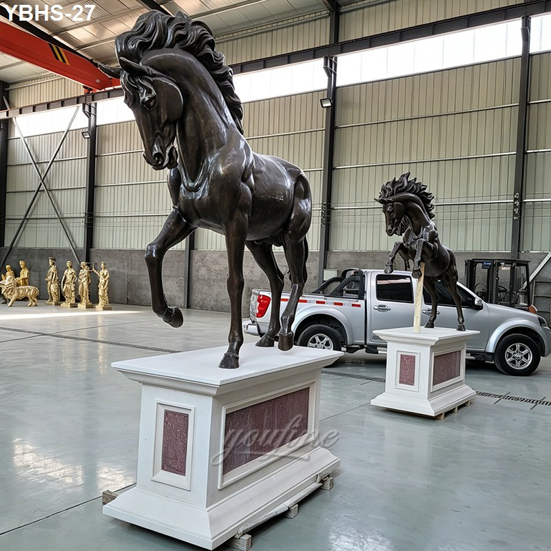 Two elegant bronze horse sculptures with detailed craftsmanship displayed on white pedestals in a spacious warehouse.
