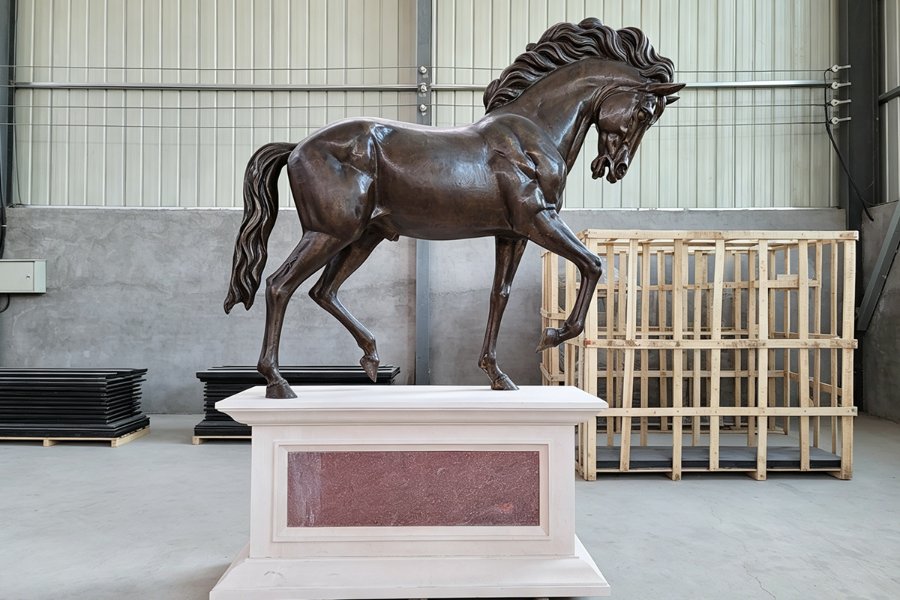 Elegant bronze standing horse sculpture with intricate detailing, mounted on a stone pedestal, placed in a large indoor workshop or gallery.