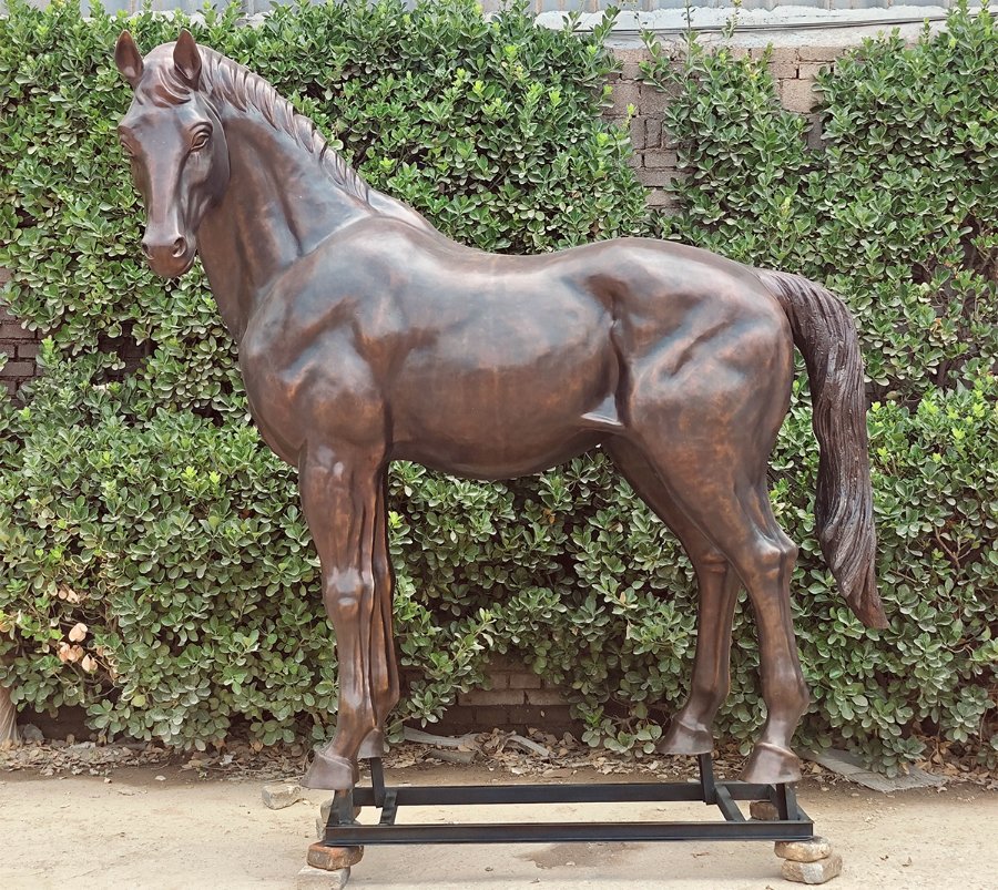 Realistic bronze horse sculpture on a metal base with intricate details, set against leafy green shrubbery in an outdoor garden.