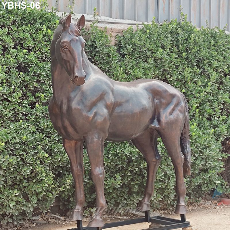 Realistic bronze horse statue standing outdoors in a landscaped garden with dense greenery and a metal fence backdrop.
