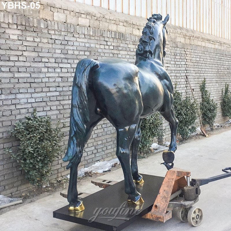 A large bronze horse sculpture with intricately detailed musculature and mane placed on a moving platform near a brick wall outdoors.