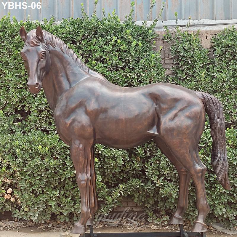 A majestic bronze horse sculpture with detailed musculature and graceful posture, displayed outdoors against green foliage and a brick wall.