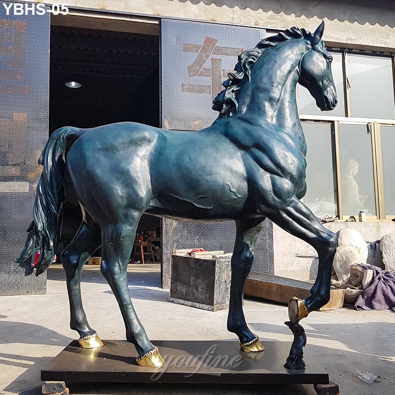 A bronze horse sculpture featuring muscular detail and gold-accent hooves, standing on a platform in a workshop setting.