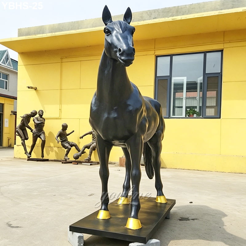 A black bronze horse statue with golden hooves displayed outdoors against a yellow wall, with smaller human sculptures visible in the background.