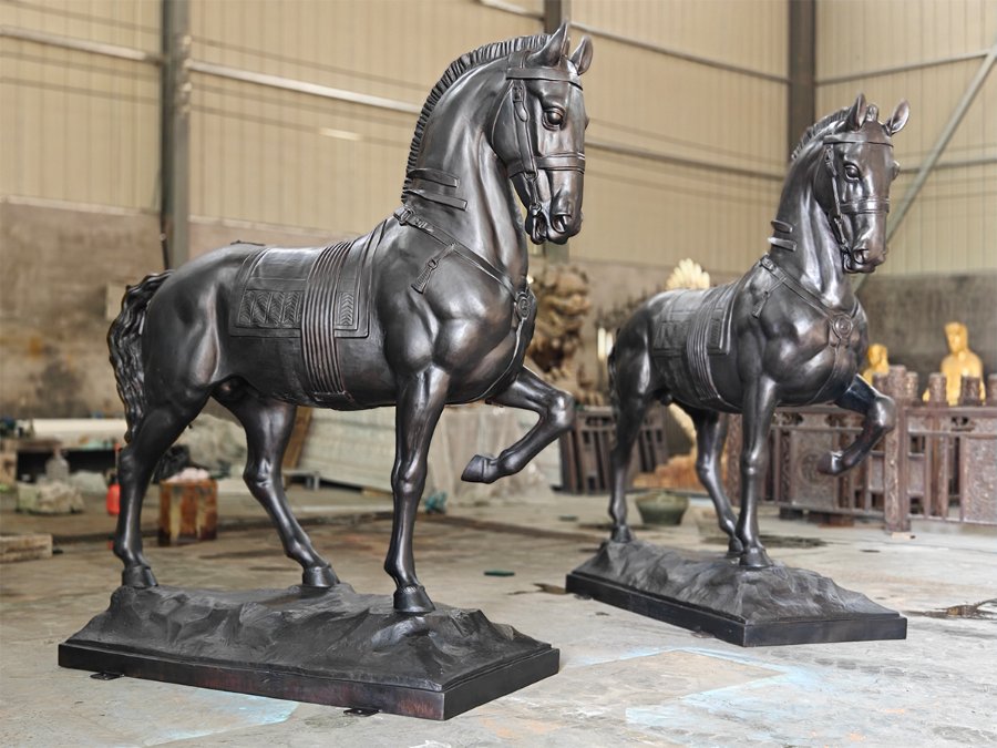 Two detailed bronze horse sculptures with dynamic poses and intricate harness designs displayed in a workshop setting.