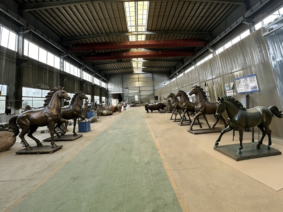 Large workshop featuring bronze horse sculptures of various designs and sizes arranged in rows. High ceiling and industrial lighting.