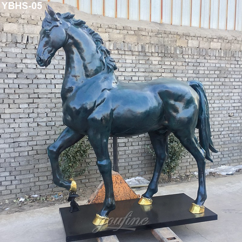 Bronze horse sculpture standing gracefully on a black base, featuring golden hooves. Positioned near a brick wall in an outdoor setting.