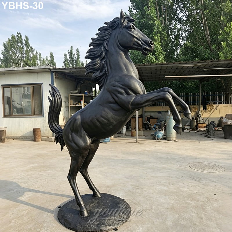 Rearing bronze horse sculpture with intricate detailing on a round base, displayed outdoors near workshop buildings and greenery.