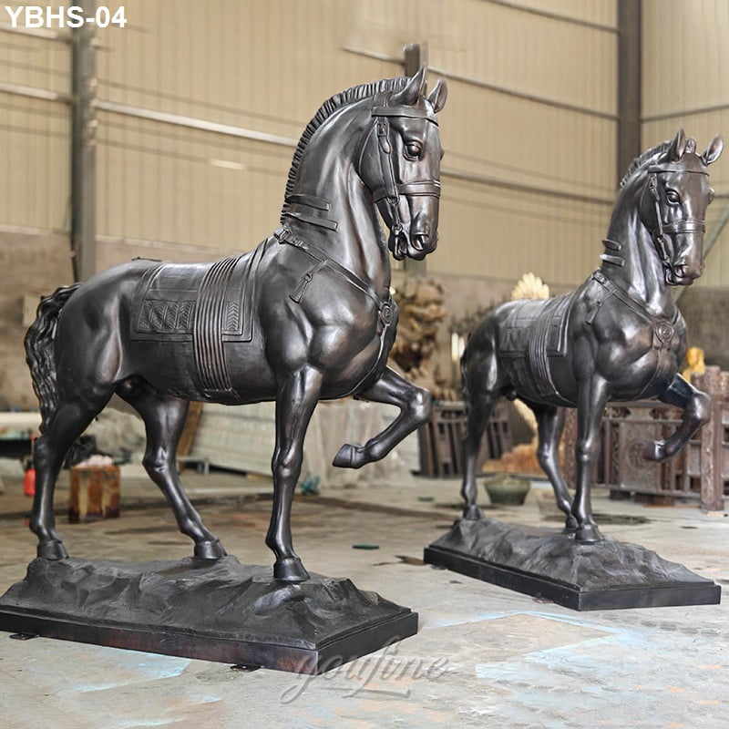Two detailed bronze horse sculptures with raised legs and ornate saddles, displayed indoors in a spacious workshop or warehouse.