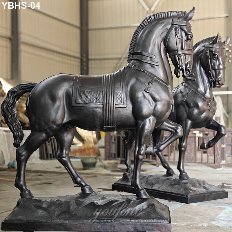 Two life size bronze horse statues with intricate saddle detailing placed on textured bases in a workshop environment.