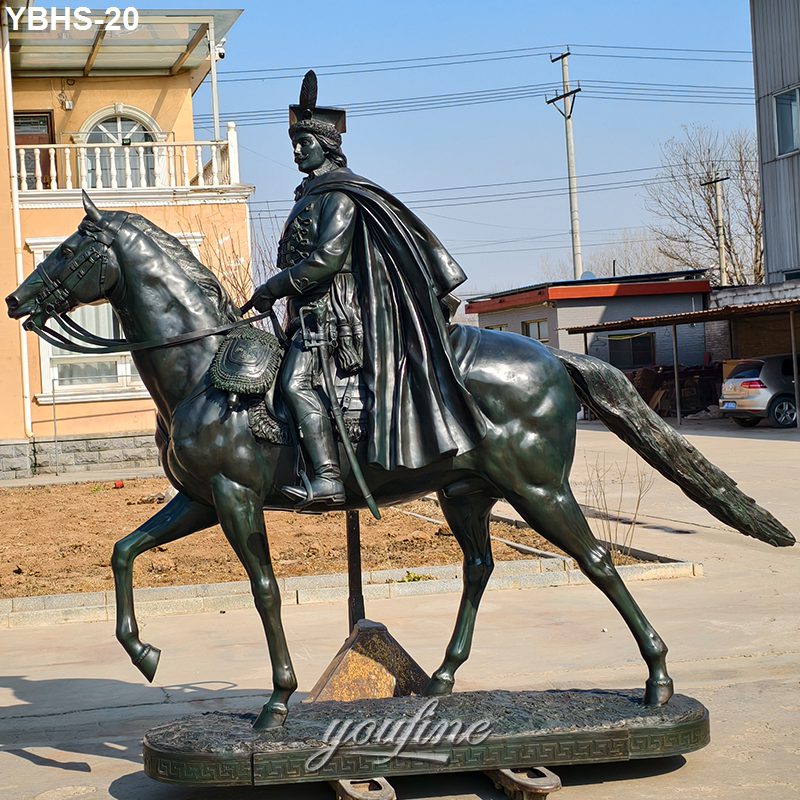 Majestic bronze horseman statue with cape and feathered hat, mounted on a pedestal in an outdoor setting near buildings.