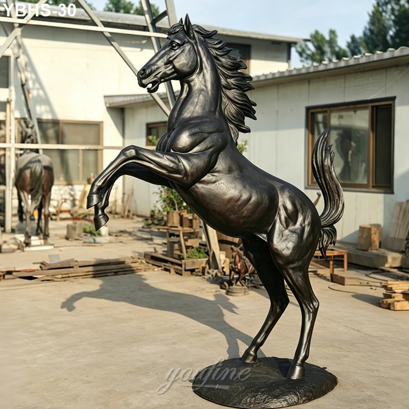 Bronze rearing horse statue with muscular details displayed outdoors in a workshop setting with sunlight highlighting its features.