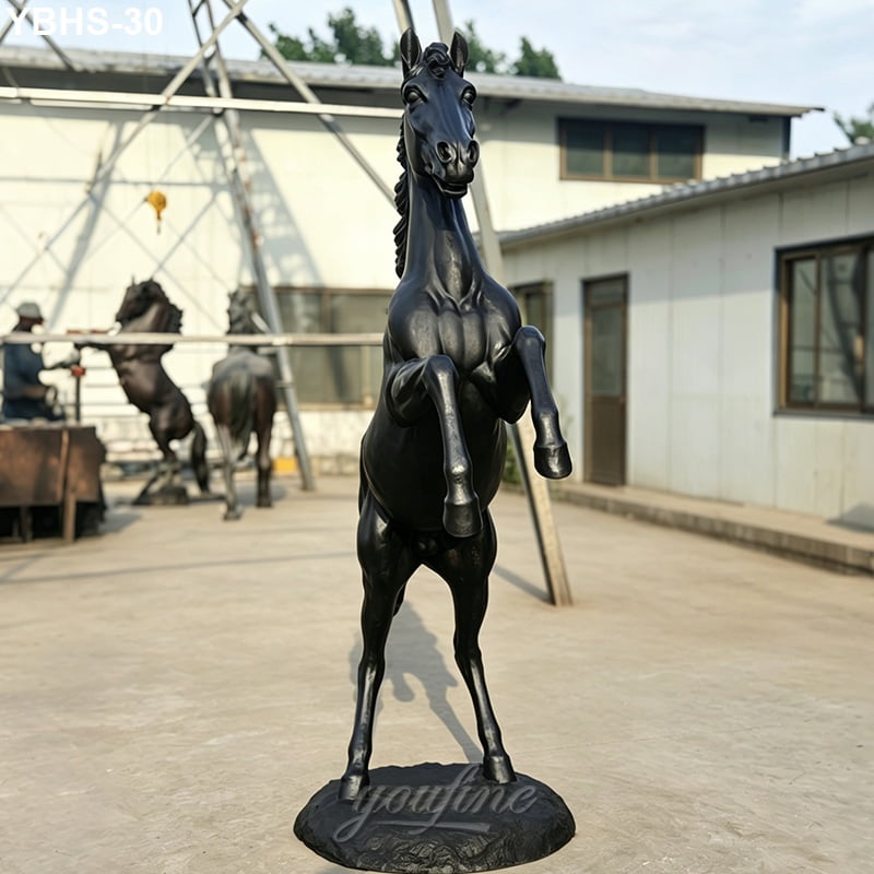 A striking bronze horse sculpture in a rearing pose placed outdoors in a workshop setting, with tools and additional sculptures visible in the background.