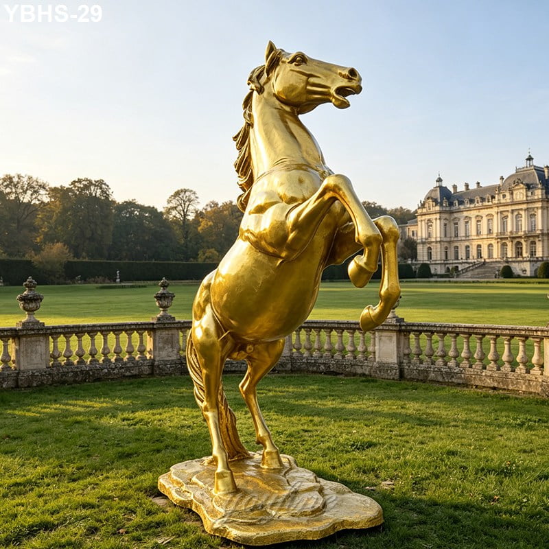A gold bronze horse statue rearing majestically on its hind legs in a lush garden with a grand castle in the background.