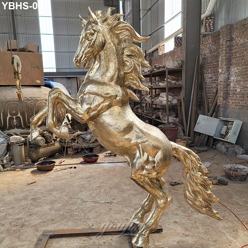 A gold bronze horse sculpture in a dynamic rearing pose, placed in an artisan workshop with tools and molds in the background.