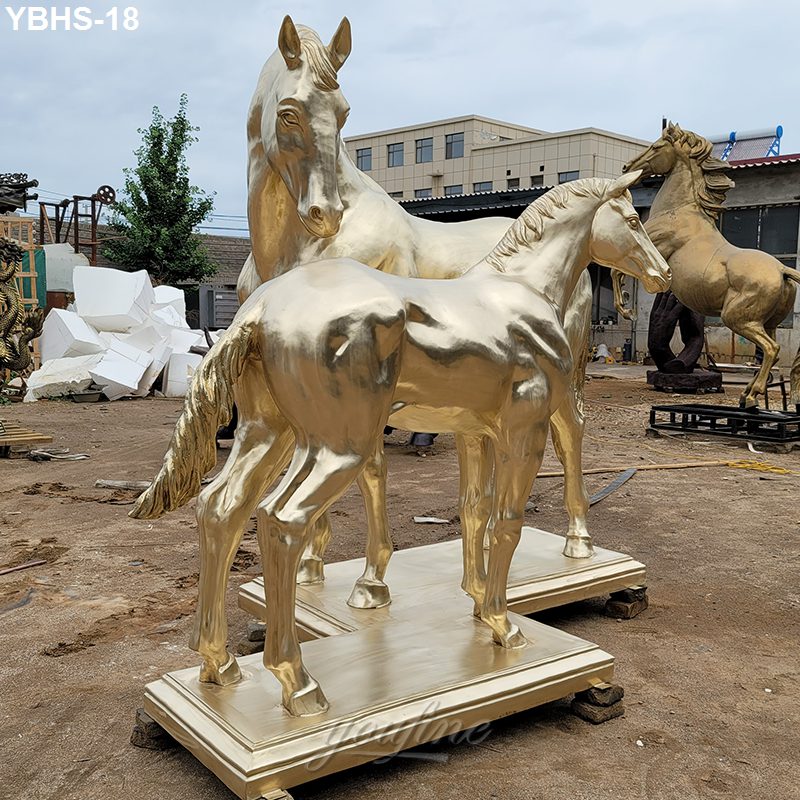 Two golden bronze horse sculptures, an adult and a foal, displayed on stone bases in an open workshop setting with other statues nearby.