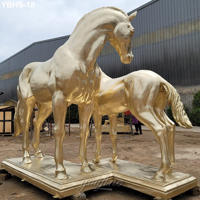 Two polished golden bronze horse sculptures with intricate detailing, standing majestically on a shared base outdoors near an industrial building.
