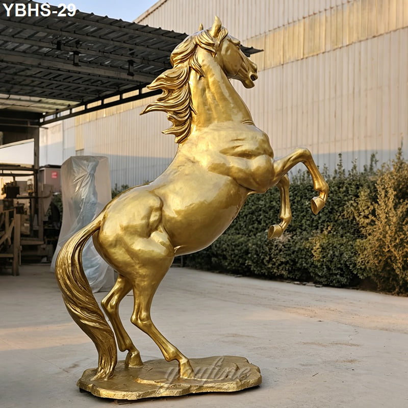 A golden bronze statue of a rearing horse with dynamic mane detailing, set outdoors near a metal structure and green plants.