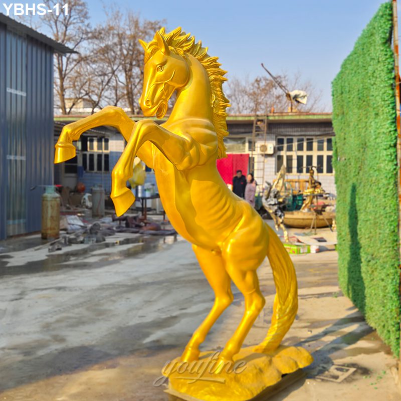 Rearing gold-painted bronze horse sculpture with detailed mane and muscular form, displayed outdoors in a workshop setting.