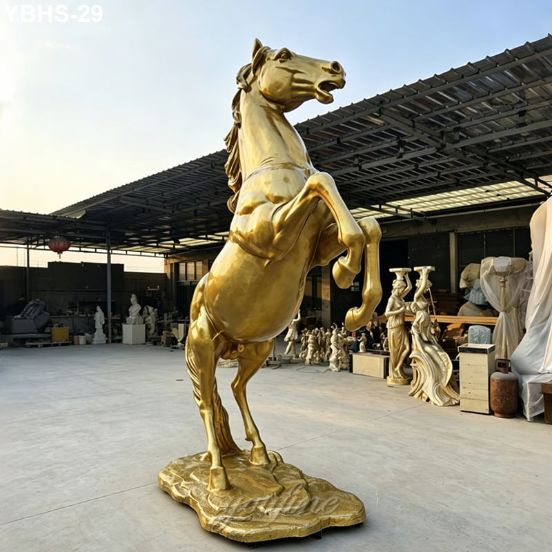 A golden bronze horse statue in a rearing position displayed in an outdoor sculpture studio with various artworks visible in the background.