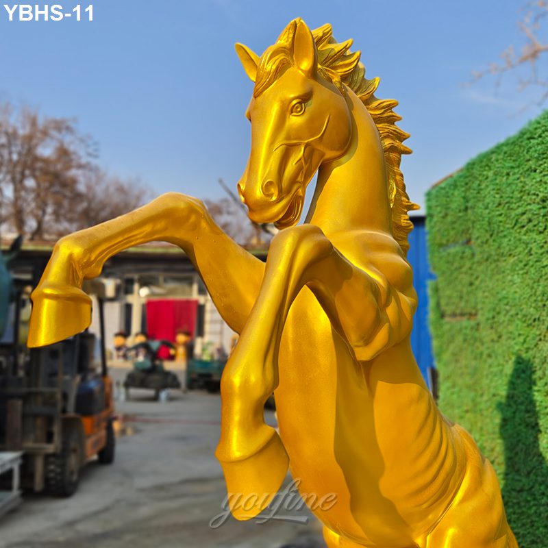 A striking golden rearing horse sculpture with realistic detailing, captured outdoors near a green hedge and workshop area.