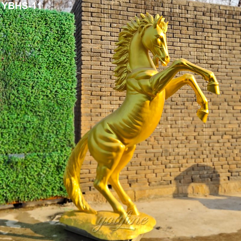A detailed golden rearing horse statue set against a brick wall and green hedge, symbolizing strength and prestige.