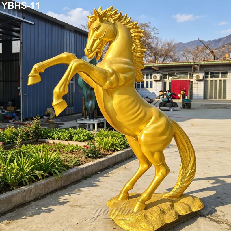 A golden rearing horse statue with a detailed mane and muscular physique placed outdoors, surrounded by plants and buildings.
