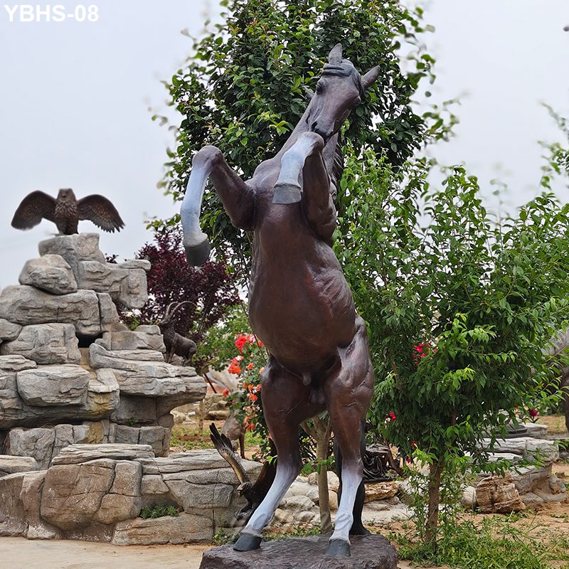 A lifelike bronze statue of a rearing horse displayed in a landscaped garden with rocks, greenery, and other sculptures.