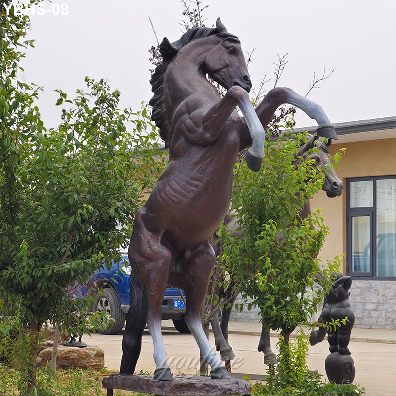 Bronze sculpture of a rearing horse with dynamic posture displayed outdoors amidst greenery and a building in the background.