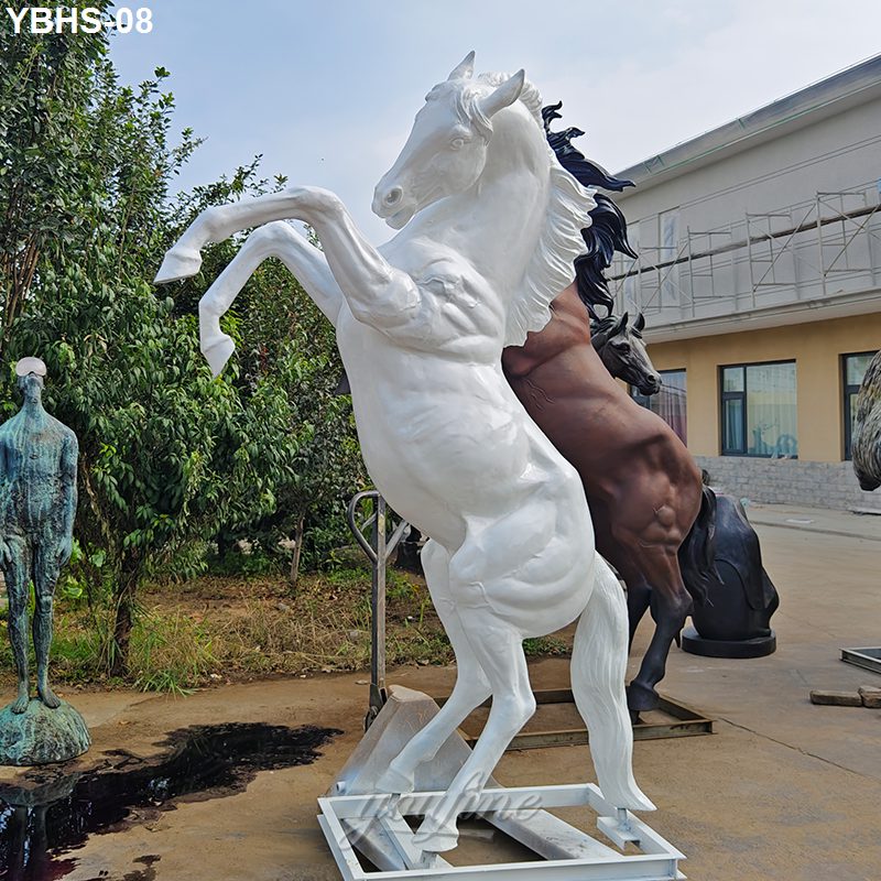A detailed white rearing horse sculpture displayed outdoors alongside other statues, with trees and a building in the background.