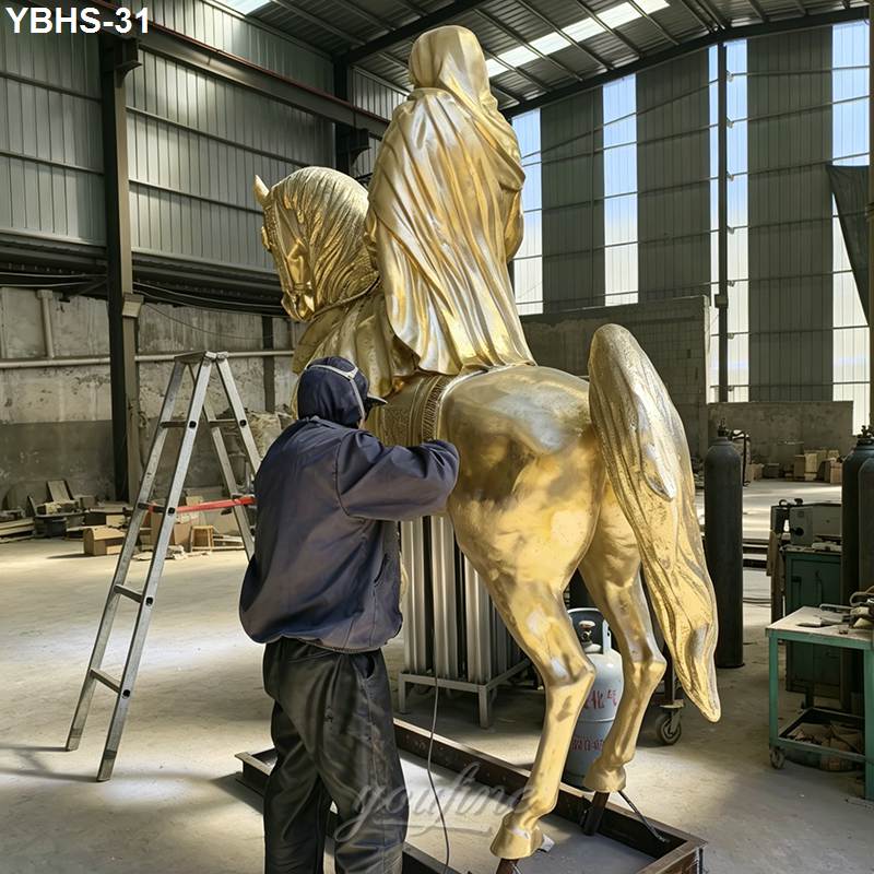 Artisan wearing a hooded jacket polishing a golden bronze horse statue with a robed rider in a workshop setting.