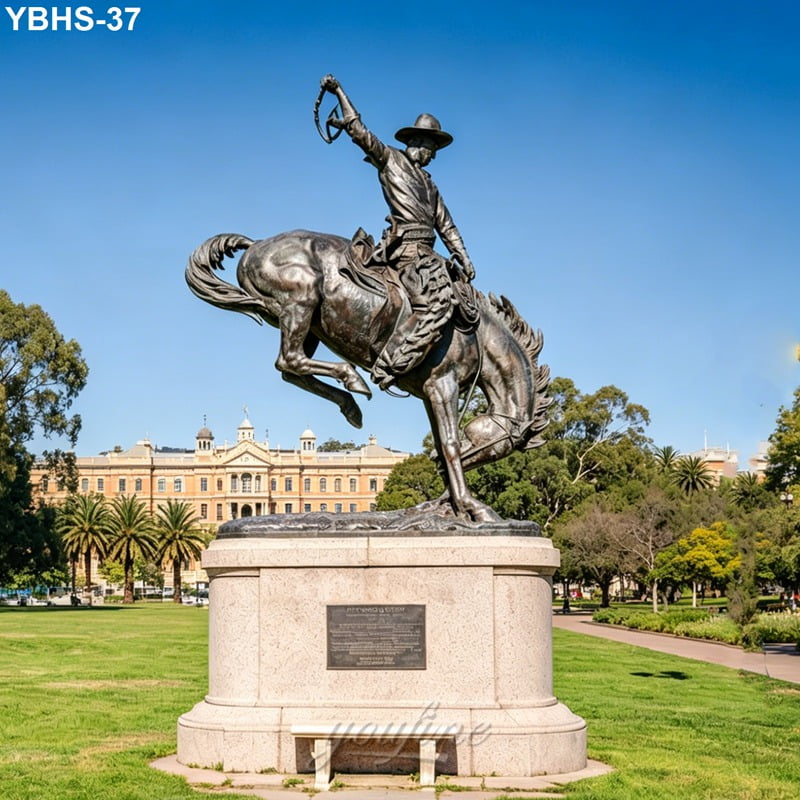 A bronze statue of a cowboy on a rearing horse, holding a whip, set on a stone pedestal in a park with a historic building in the background.