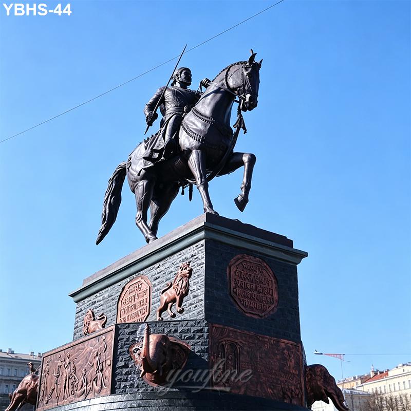 Bronze equestrian statue of a warrior holding a sword, on a detailed pedestal adorned with relief art and animal symbols in an outdoor setting.
