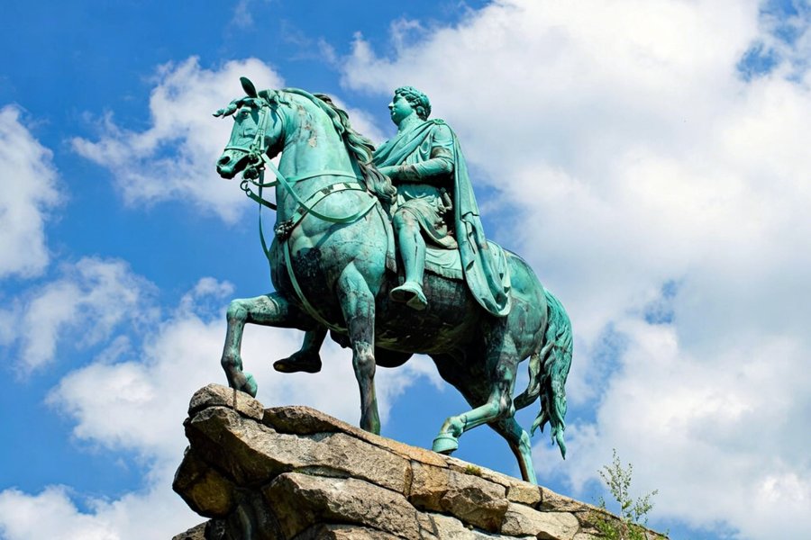 An ornate bronze equestrian statue of a figure on horseback atop a stone base, set against a bright blue sky with clouds.