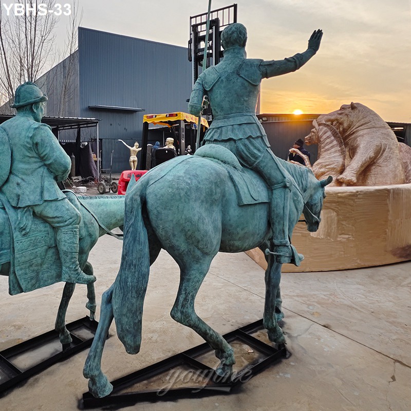 Two bronze equestrian statues depicting historical riders viewed from the backside, displayed outdoors with other sculptures near a workshop at sunset.